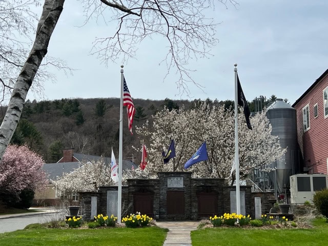 a stone memorial with six flags stands among blooming trees and daffodils, with buildings and wooded hills in the background on a cloudy day.