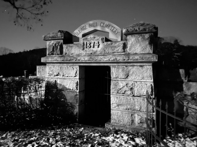 black and white photo of a stone mausoleum at village hill cemetery, dated 1874, with an open doorway and surrounding iron fence. fallen leaves cover the ground.