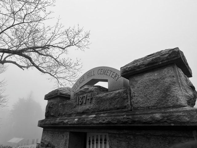 black and white photo of the stone entrance to village hill cemetery, with "1874" engraved on it; bare tree branches and fog are visible in the background.