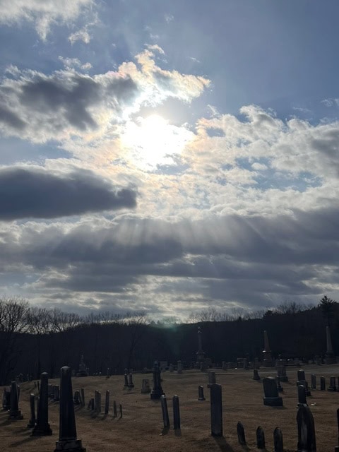 the sun breaks through scattered clouds, casting rays of light over a cemetery with numerous upright and leaning gravestones.