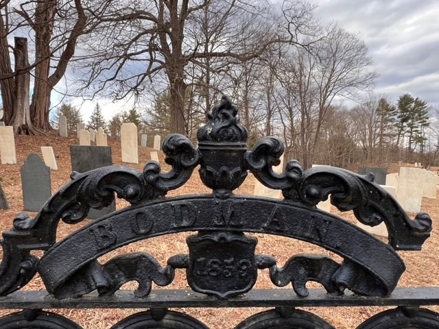 black wrought iron gate with the inscription "bodman 1839" in front of an old cemetery with weathered tombstones and leafless trees.