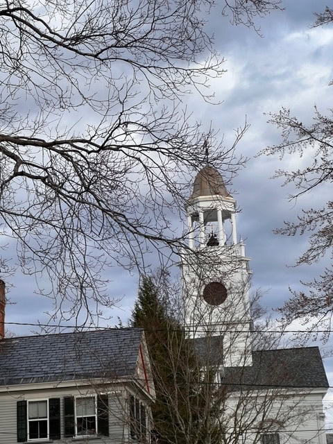 white church steeple with a clock rises above nearby buildings, framed by bare tree branches under a cloudy sky.