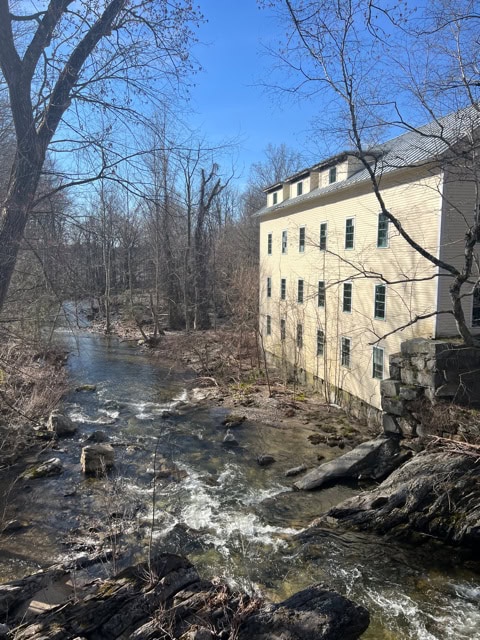 a cream colored building with many windows sits beside a rocky stream surrounded by leafless trees under a clear blue sky.