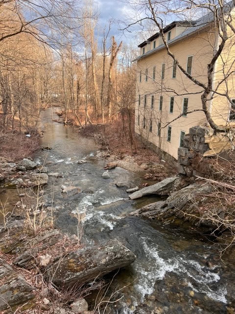a stream with small rapids flows past a large, beige building with many windows; leafless trees line both sides under a partly cloudy sky.