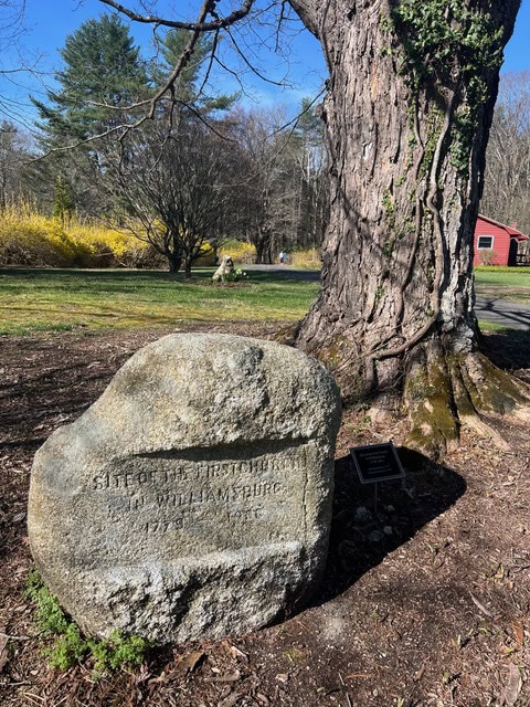 large rock with inscription marking the site of the first home in williamsburg, 1735; situated near a tree in a grassy park with a red building in the background.