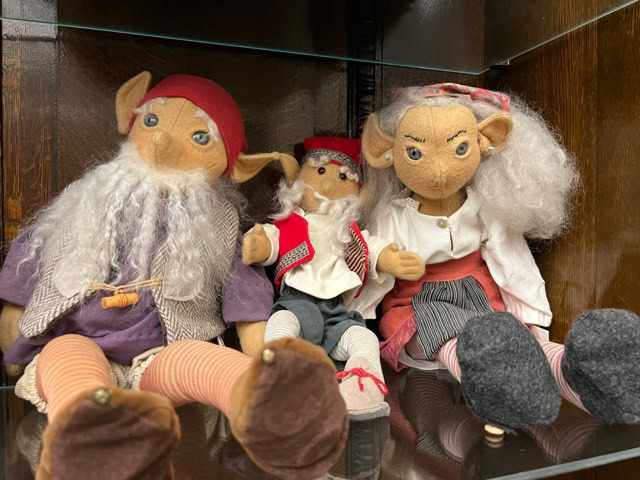 three handmade cloth dolls with pointy ears, dressed in folk style clothing, are seated on a wooden shelf with glass panels.