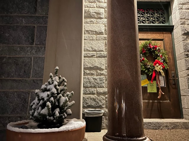 a potted evergreen dusted with snow sits near a column in front of a stone building, with a door decorated with a holiday wreath and papers attached.