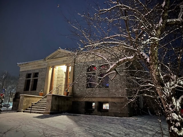 a historic stone building with columns and illuminated entrance stands beside a snow covered tree at night.
