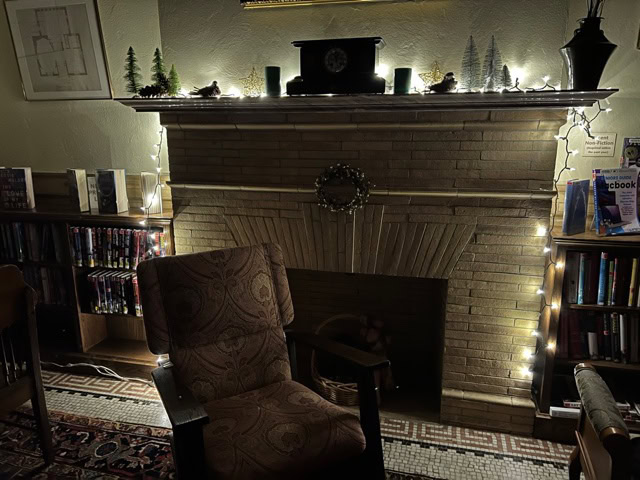 a cozy armchair sits in front of a fireplace decorated with string lights, small pine tree figurines, and bookshelves on either side.