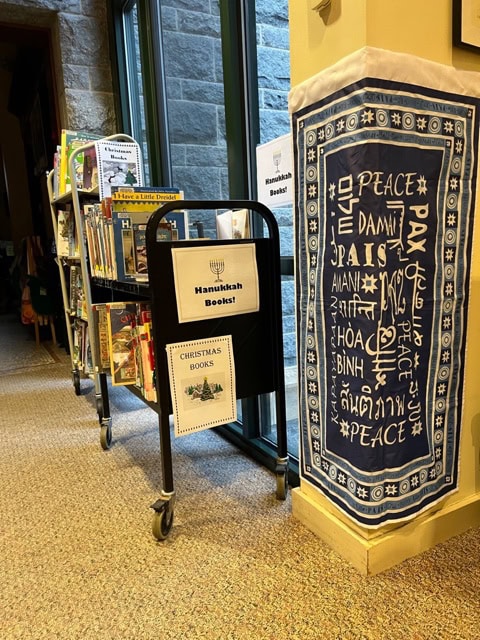 library book carts labeled "hanukkah books" and "christmas books" stand next to a wall hanging with the word "peace" in multiple languages.
