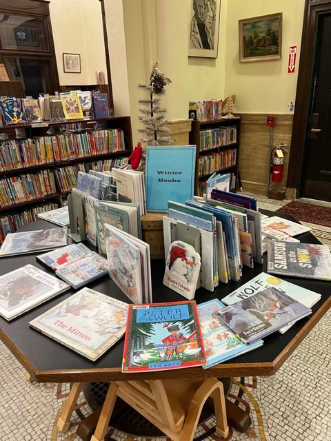 a library display table with a selection of winter themed children's books arranged around a small decorative tree, with shelves of more books in the background.