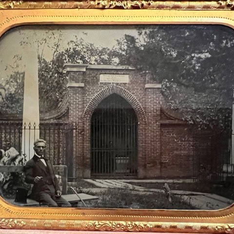 a black and white photograph in a gold frame shows a man seated outside a gated brick tomb entrance with iron fencing and trees in the background.