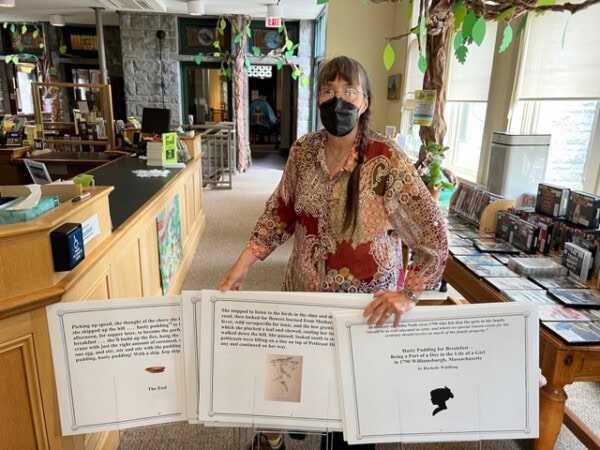 a person wearing a patterned shirt and face mask stands in a library, holding large display boards with text and images on them.