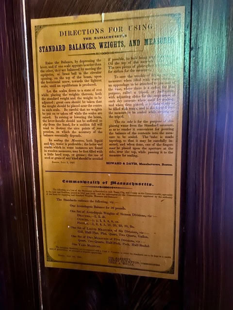 a yellowed sign on a wooden wall displays instructions for using massachusetts standard balances, weights, and measures, with additional legal information below.