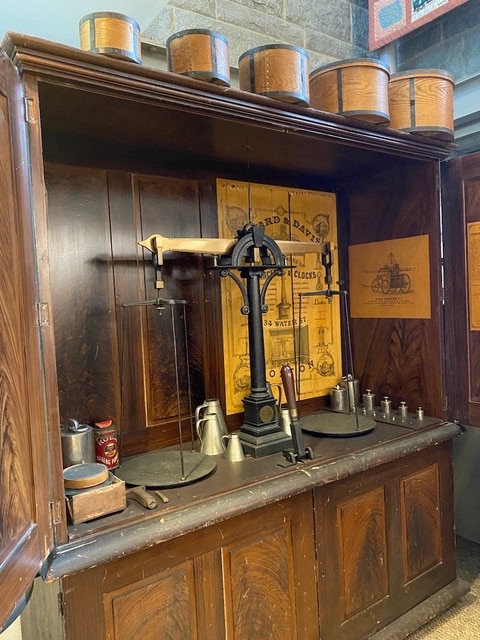 antique balance scale and weights displayed in a wooden cabinet, with tins on top and vintage posters on the back wall.
