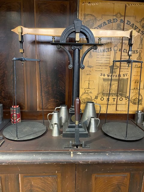 an antique balance scale with metal weights and measures sits on a wooden counter; a vintage advertisement is visible in the background.