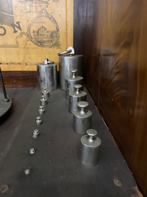 a row of metal calibration weights of varying sizes is lined up on a dark wooden surface, with a vintage illustration partially visible in the background.