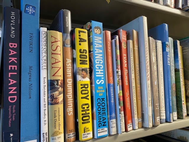 a row of cookbooks, including titles on baking, asian cuisine, korean cooking, and dumplings, arranged upright on a library shelf.
