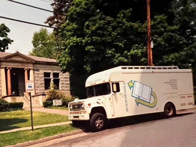 a white bookmobile is parked on a street in front of a small stone library building, surrounded by trees and greenery.