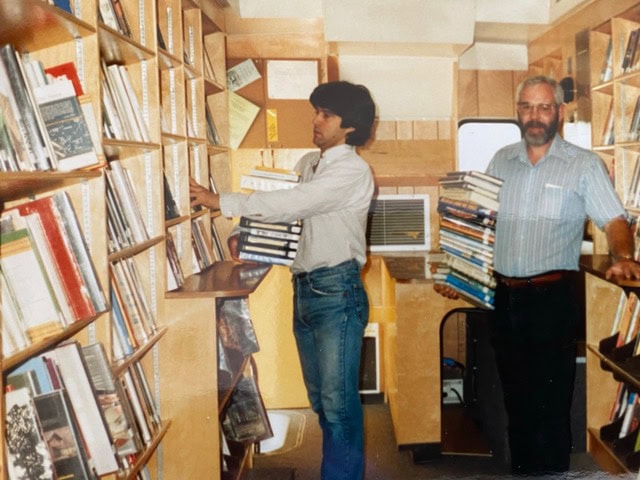 two men organize and shelve books inside a small, wood paneled library or bookmobile, each holding a stack of books. shelves are filled with various books.