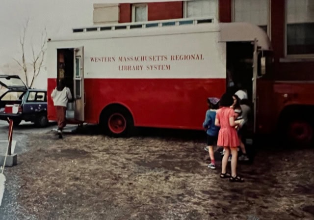 a red and white bookmobile labeled "western massachusetts regional library system" is parked beside a building as several children and a woman enter or stand nearby.