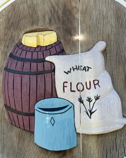 wooden relief carving of a barrel, a blue container, and a flour sack labeled "wheat flour" with wheat drawings, mounted on a wood background.