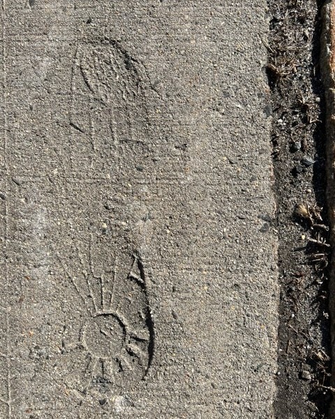 a faint shoe print with a sunburst pattern on the sole is visible on a rough concrete sidewalk.