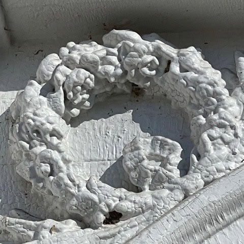 close up of a textured, white painted wreath in relief on a cracked and weathered surface, showing peeling paint and shadow details.