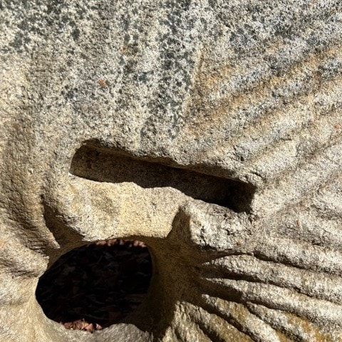 a close up view of a weathered stone surface with a carved hole and grooved lines radiating outward, illuminated by sunlight.