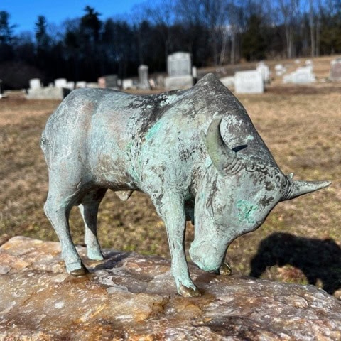 a small bronze bull statue stands on a stone in a cemetery, with gravestones and trees visible in the background.