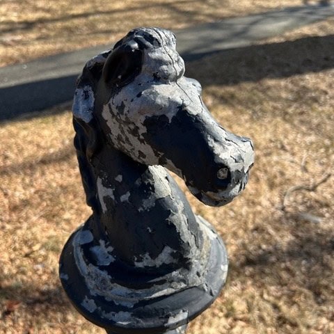a weathered black horse head hitching post with peeling paint stands outdoors on dry grass with a paved path in the background.