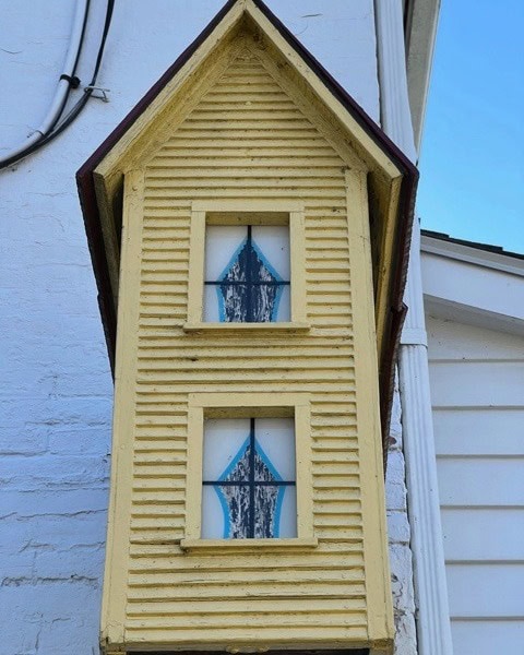 a small, yellow, house shaped structure with two blue trimmed windows is mounted against the side of a white building.