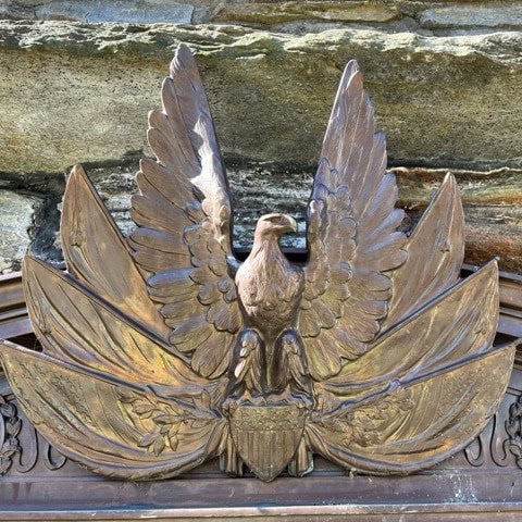 a bronze eagle sculpture with outstretched wings sits atop a shield and laurel leaves, mounted against a stone wall.