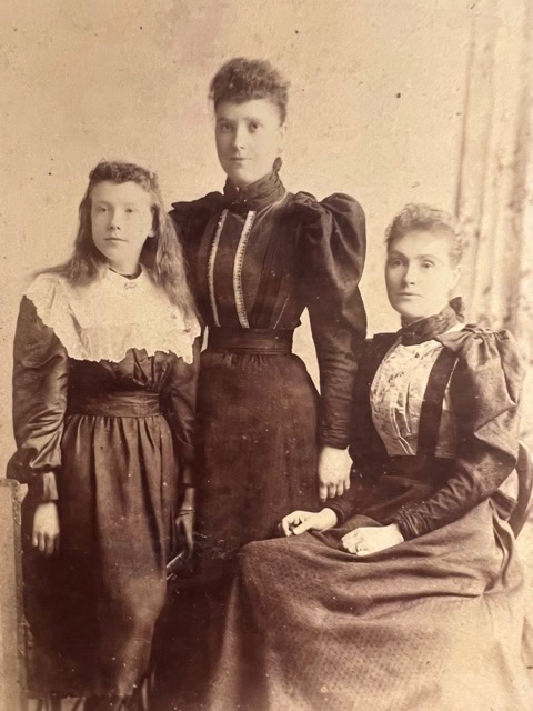 three women in late 19th century or early 20th century dresses pose for a formal sepia toned portrait, with two standing and one seated.