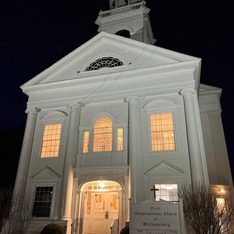 white church building illuminated at night, with lit windows and a sign reading “first congregational church of williamsburg” in front of the entrance.