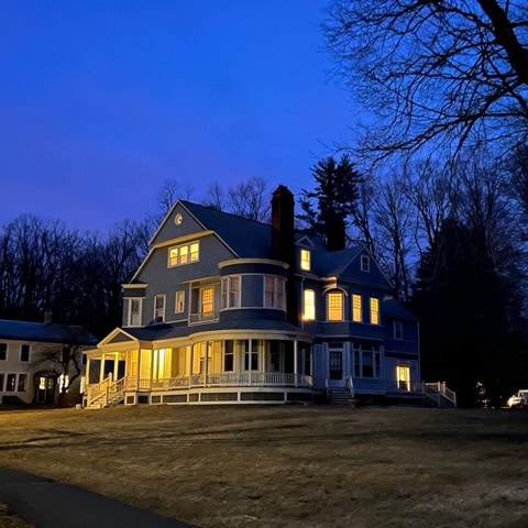 a large, two story house with lit windows stands on a lawn at dusk, surrounded by bare trees under a deep blue sky.