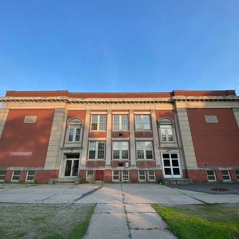 a front view of an old, red brick school building with large windows, double doors, and a faded concrete walkway leading to the entrance.