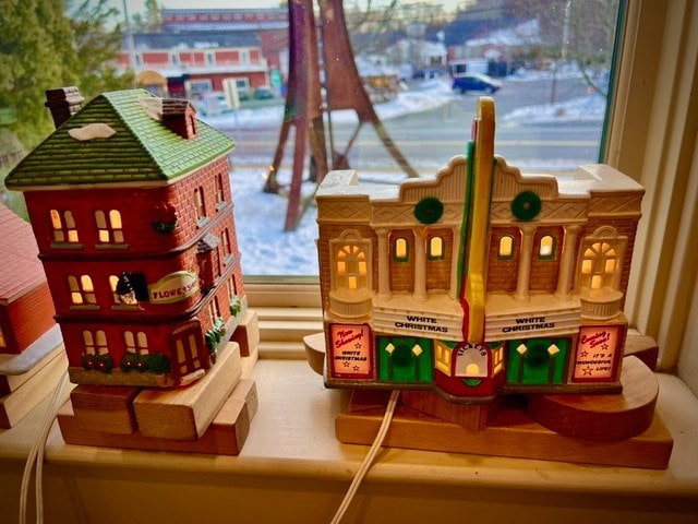 two miniature, illuminated ceramic buildings—a flower shop and a theater—sit on a windowsill with a snowy outdoor scene visible through the window.