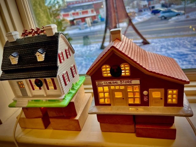two illuminated miniature ceramic buildings, a house and a general store, are displayed on a windowsill with a snowy street scene visible outside.