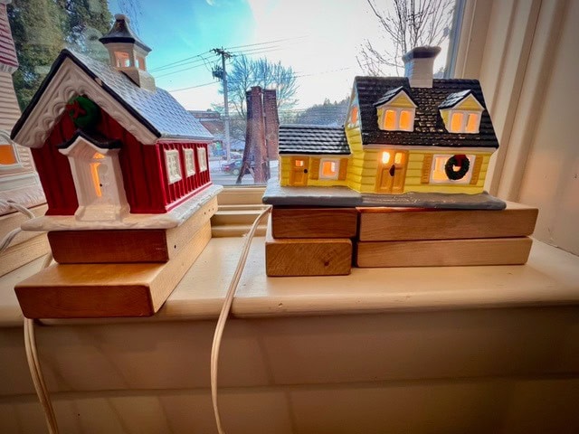 two decorative ceramic buildings, one red and one yellow, sit on wooden blocks by a window with cords plugged in, their windows softly lit from within.