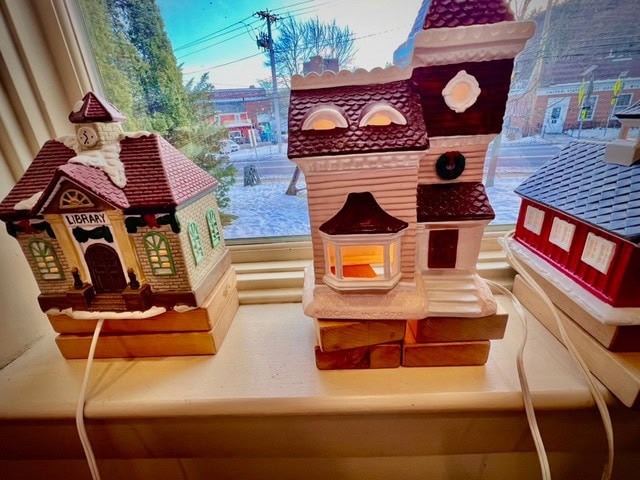 three miniature ceramic buildings, including a library and two houses, are displayed on a windowsill with cords attached, against a backdrop of a snowy street outside.
