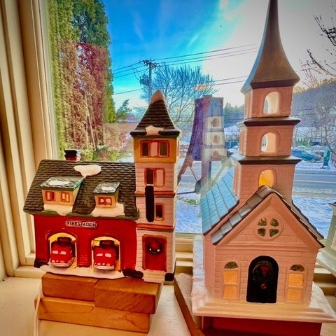 two ceramic buildings, a fire station and a church, are displayed on a windowsill with a winter scene visible outside.