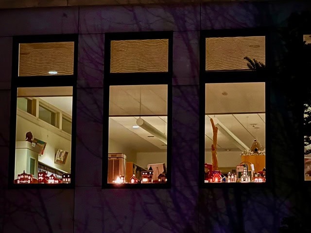 three large windows reveal a brightly lit indoor space with shelves, books, and various decorations visible at night.