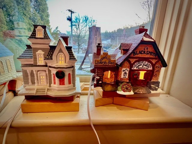 two miniature ceramic buildings, a victorian style house and a blacksmith shop, are displayed on a windowsill with trees and power lines visible outside.