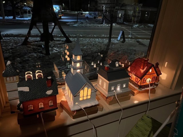 small ceramic houses with illuminated windows are displayed on a windowsill at night, with snow visible outside and buildings across the street.