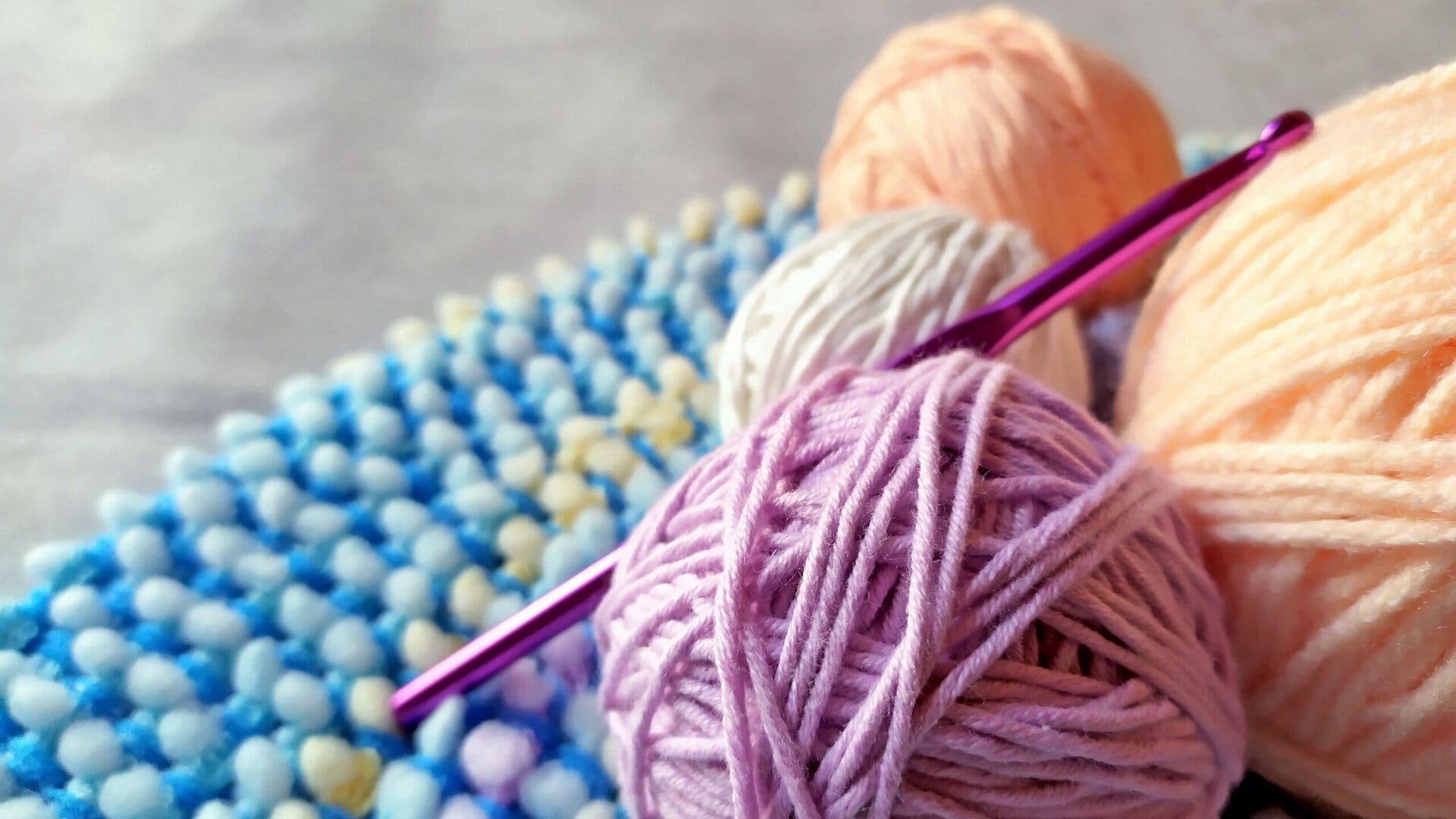three balls of yarn and a purple crochet hook rest on a blue textured mat with yellow and white dots.